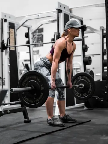 A woman wearing a cap and workout clothes lifts a barbell while performing a deadlift in a gym.