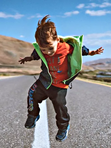 A young child in colorful clothes joyfully jumps on a deserted road surrounded by hills under a bright blue sky.Pediatric Chiropractic