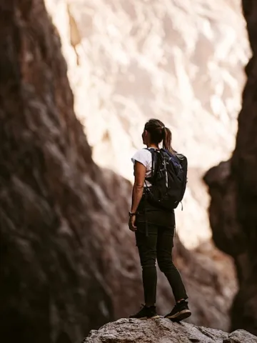 A woman wearing a backpack stands on a rock, gazing up at towering canyon walls in a sunlit mountain pass.