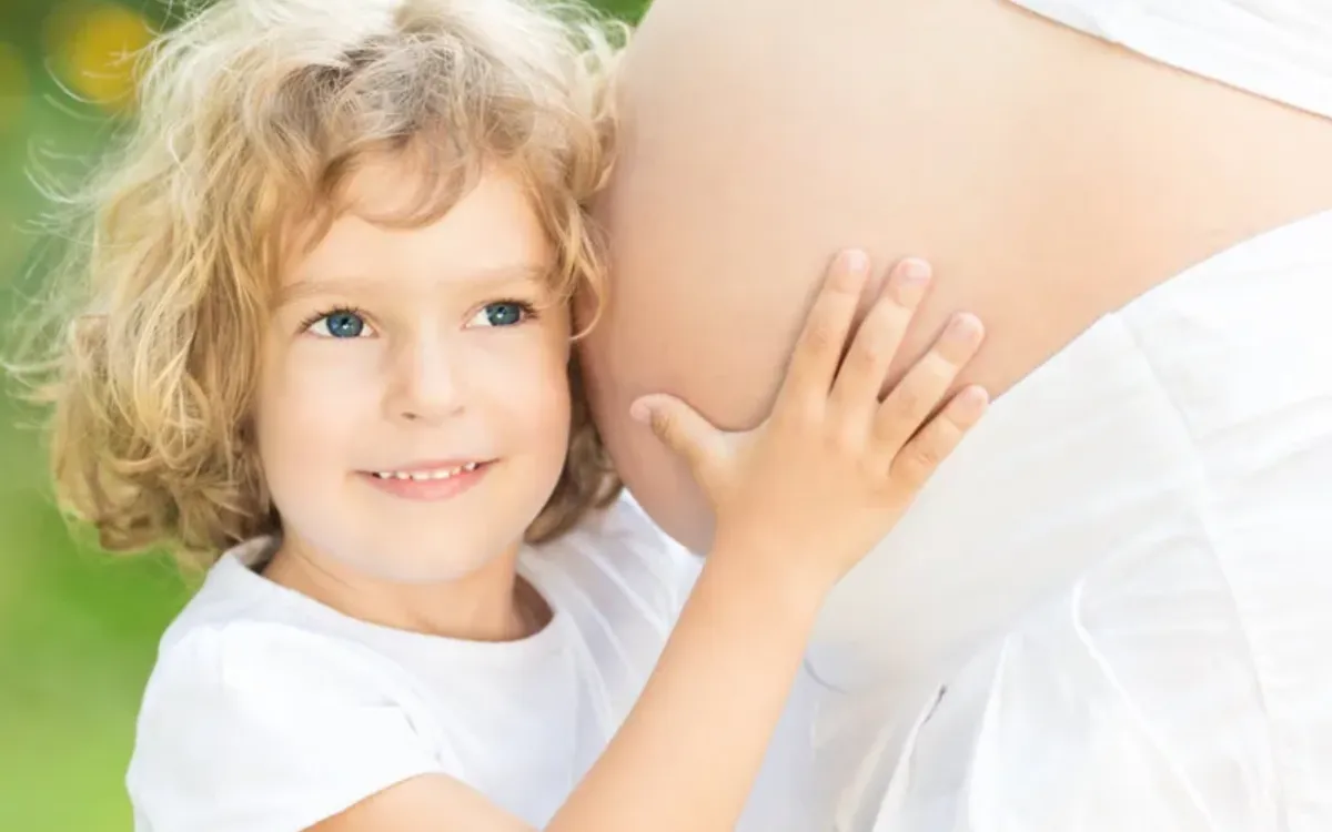 A young child with curly blonde hair smiles while gently placing a hand on a pregnant woman's belly.