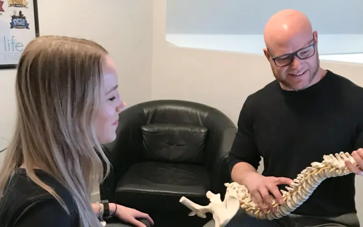 Dr. Richards holds and explains a model of the human spine to a smiling female patient during a chiropractic consultation.