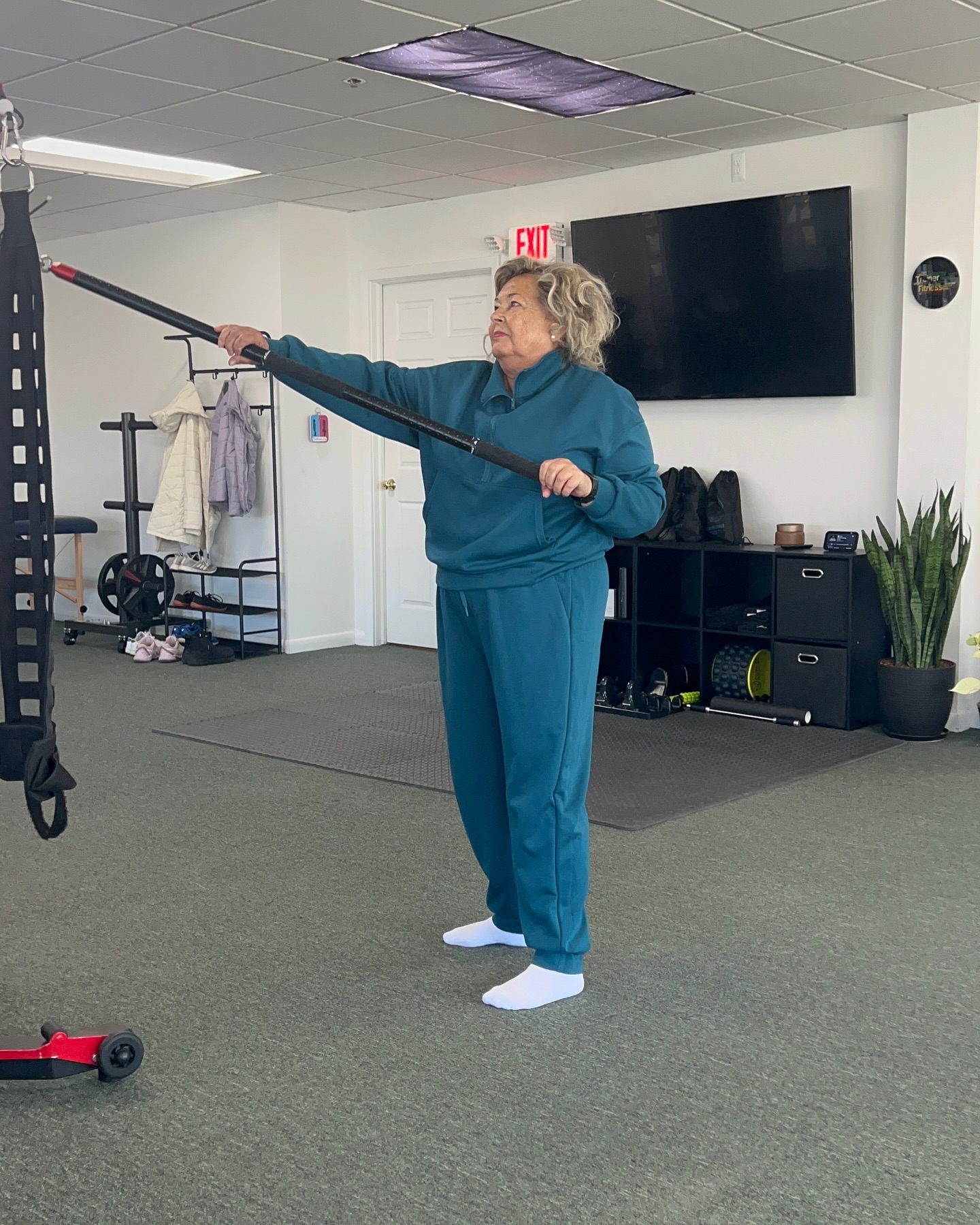Person at a personal trainer holding a long black exercise bar while standing in a teal tracksuit and white socks, with workout equipment and a wall-mounted TV in the background.