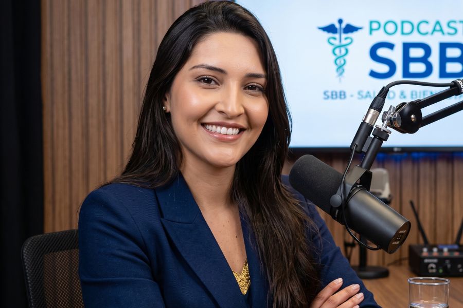 Alt text: Un retrato fotográfico profesional de la Dra. Daniela Garcés, una mujer de cabello oscuro, sentada en un estudio de podcast y sonriendo a la cámara con los brazos cruzados sobre una mesa de madera. Viste un blazer azul marino y joyas doradas. Delante de ella hay un micrófono profesional y un vaso de agua. Al fondo, una pantalla de televisión muestra un logo médico con la inscripción "PODCAST SBB - SALUD & BIENESTAR" sobre una pared panelada de madera.