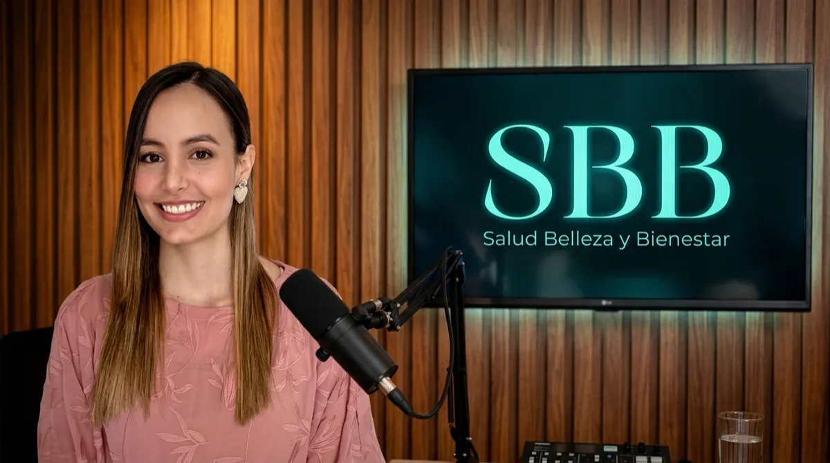 Una mujer joven sonriente con cabello largo, castaño claro con mechas doradas y partido al lado. Lleva puesta una blusa rosa de manga larga con un delicado patrón de hojas bordadas o estampadas y un arete en forma de corazón. En el fondo de la imagen generada, se encuentra en un estudio de podcast sobre una pared de paneles de madera vertical cálida, con un micrófono profesional de mesa frente a ella y una pantalla al fondo que muestra el logo "SBB - Salud Belleza y Bienestar