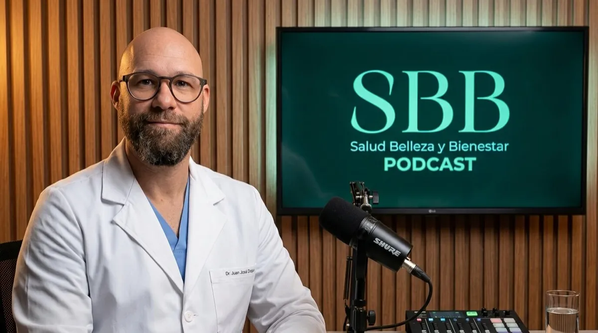 Un hombre  con barba  corta, lentes redondos de pasta oscura y expresión sonriente y profesional. Viste una bata médica blanca sobre un uniforme clínico  de color azul claro, llevando una etiqueta de identificación en el bolsillo con el nombre "Dr. Juan José Duque". Se encuentra en el lado izquierdo del encuadre dentro de un estudio de podcast con pared de listones de madera de tono cálido. Frente a él hay un micrófono profesional marca Shure sobre un brazo articulado y una mesa con consola de audio y un vaso de agua; al fondo, una pantalla muestra el logo brillante del podcast "SBB - Salud Belleza y Bienestar PODCAST" sobre un fondo verde oscuro