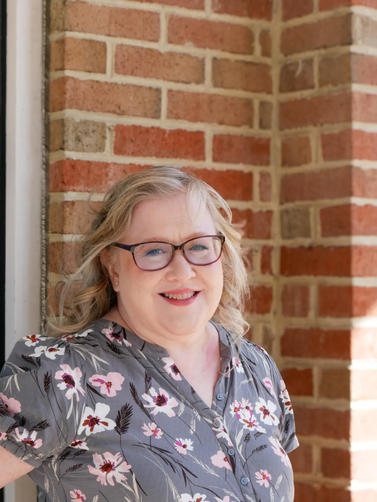 Picture of a blonde woman with glasses wearing a green floral shirt in front of a brick wall