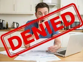 A man sitting at a table with paperwork and a laptop looks surprised; the word DENIED is stamped across the image in red.