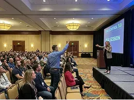 A woman speaks onstage to a large seated audience in a conference room; one attendee stands and raises his hand to ask a question.