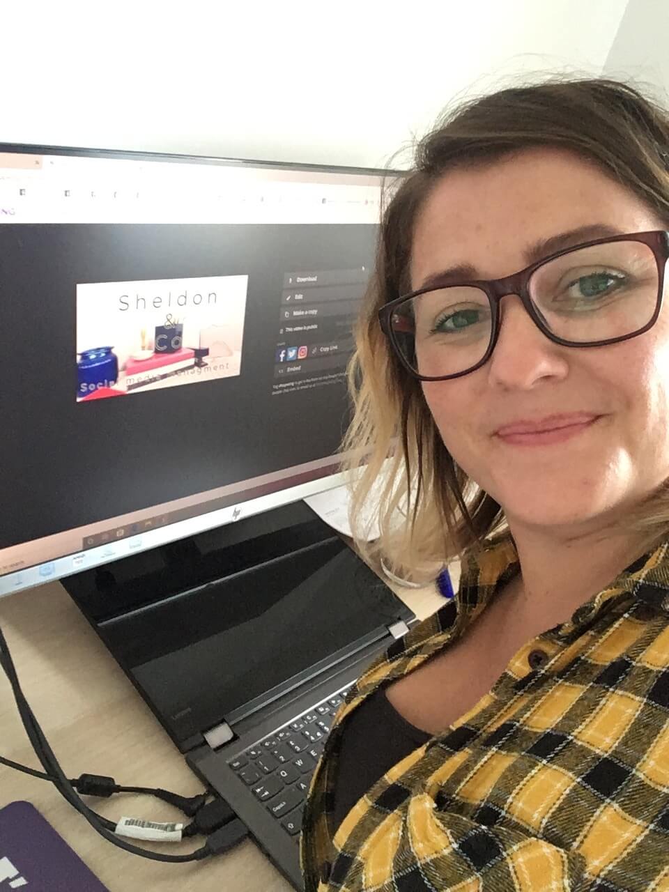 Shelly Terry, founder of Sheldon Social, smiling at the camera in her workspace with a laptop and desktop monitor visible behind her showing the Sheldon Social brand, wearing glasses and a yellow checked shirt.