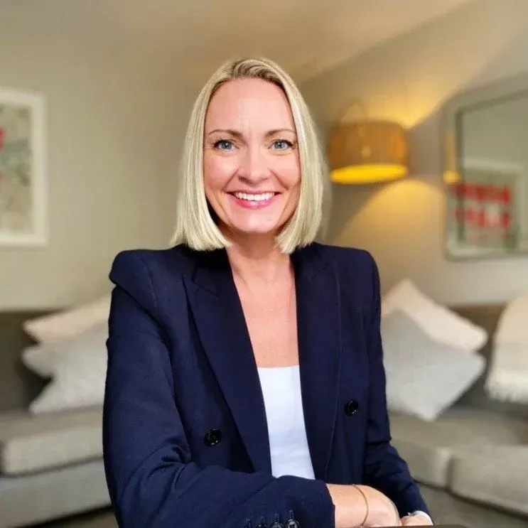 A smiling woman with a blonde bob, wearing a navy blazer, photographed in a stylish interior setting — a founder working with Sheldon Social on thought leadership content and brand voice.