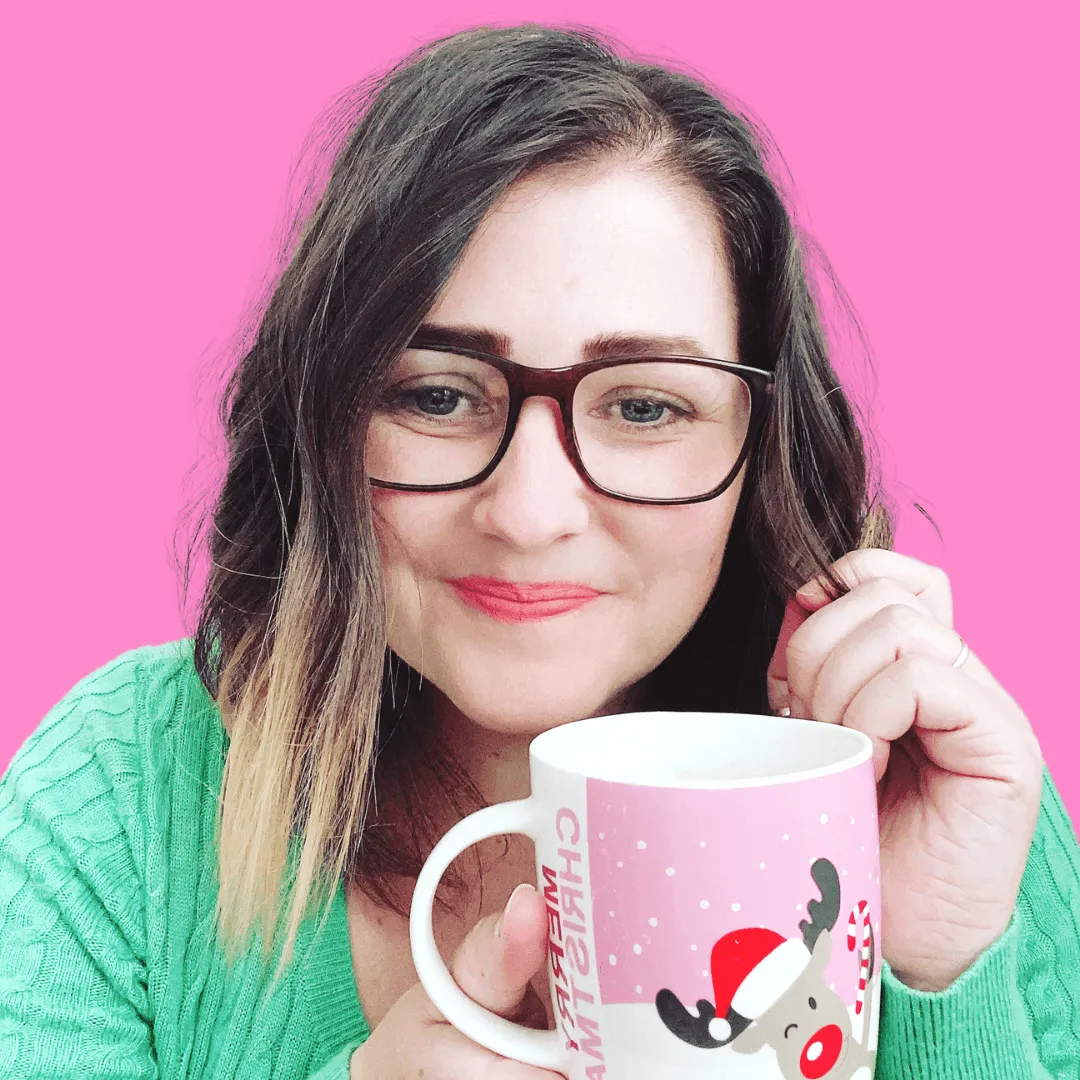 Shelly Terry, founder of Sheldon Social, smiling at the camera holding a Christmas mug, wearing glasses and a green knit jumper against a bright pink background — content strategist and visibility consultant helping founders and brands shape how they're understood online.