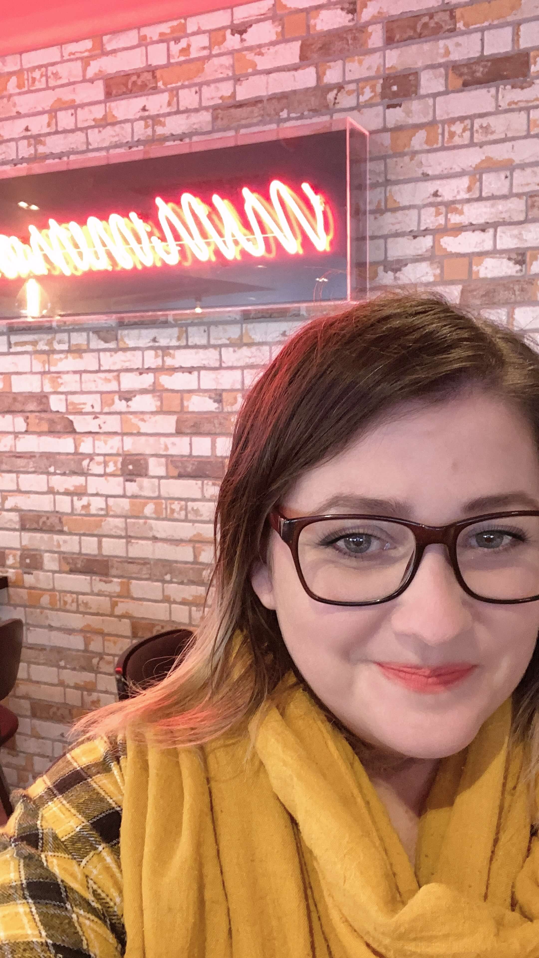 Shelly Terry, founder of Sheldon Social, smiling in a casual setting with a warm brick wall and neon sign in the background, wearing glasses and a yellow scarf — content strategist and visibility consultant helping founders and brands shape how they're understood online.