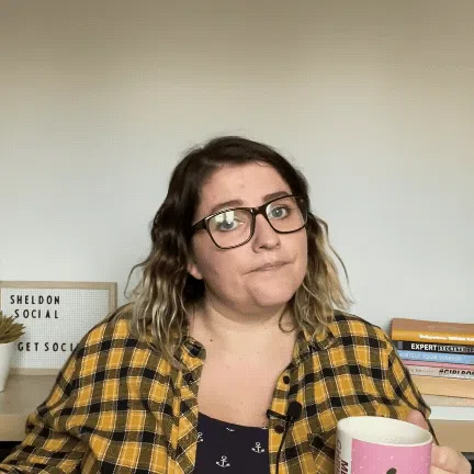 Shelly Terry, founder of Sheldon Social, holding a mug at her desk with a Sheldon Social letterboard sign and books visible in the background, wearing glasses and a yellow checked shirt — content strategist and visibility consultant helping founders and brands shape how they're understood online.