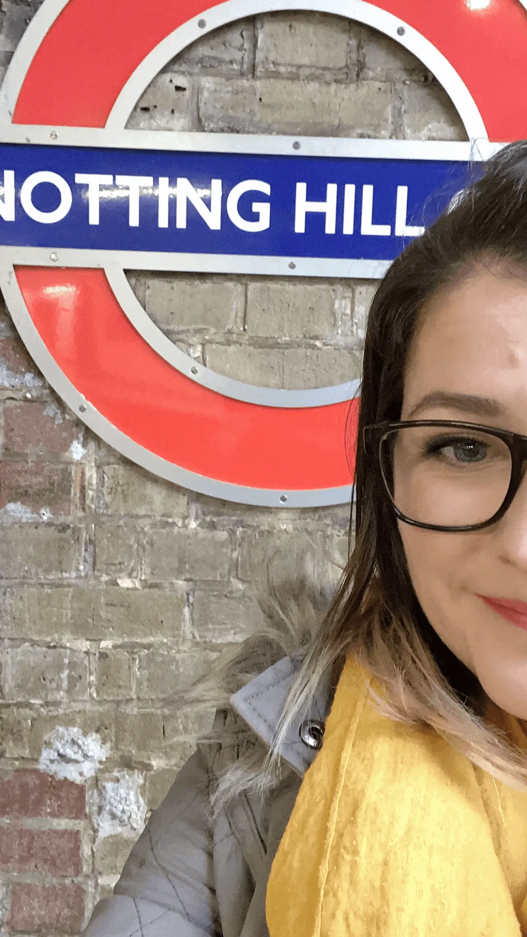 Shelly Terry, founder of Sheldon Social, wearing glasses and a yellow jumper, photographed in front of the Notting Hill London Underground roundel sign — UK-based content strategist and visibility consultant helping founders and brands shape how they're understood online.
