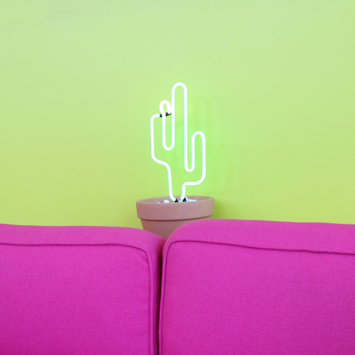 A neon cactus light in a terracotta pot placed between two bright pink sofa cushions against a yellow-green wall — used to illustrate a Sheldon Social article on content ecosystems and sustainable content systems.