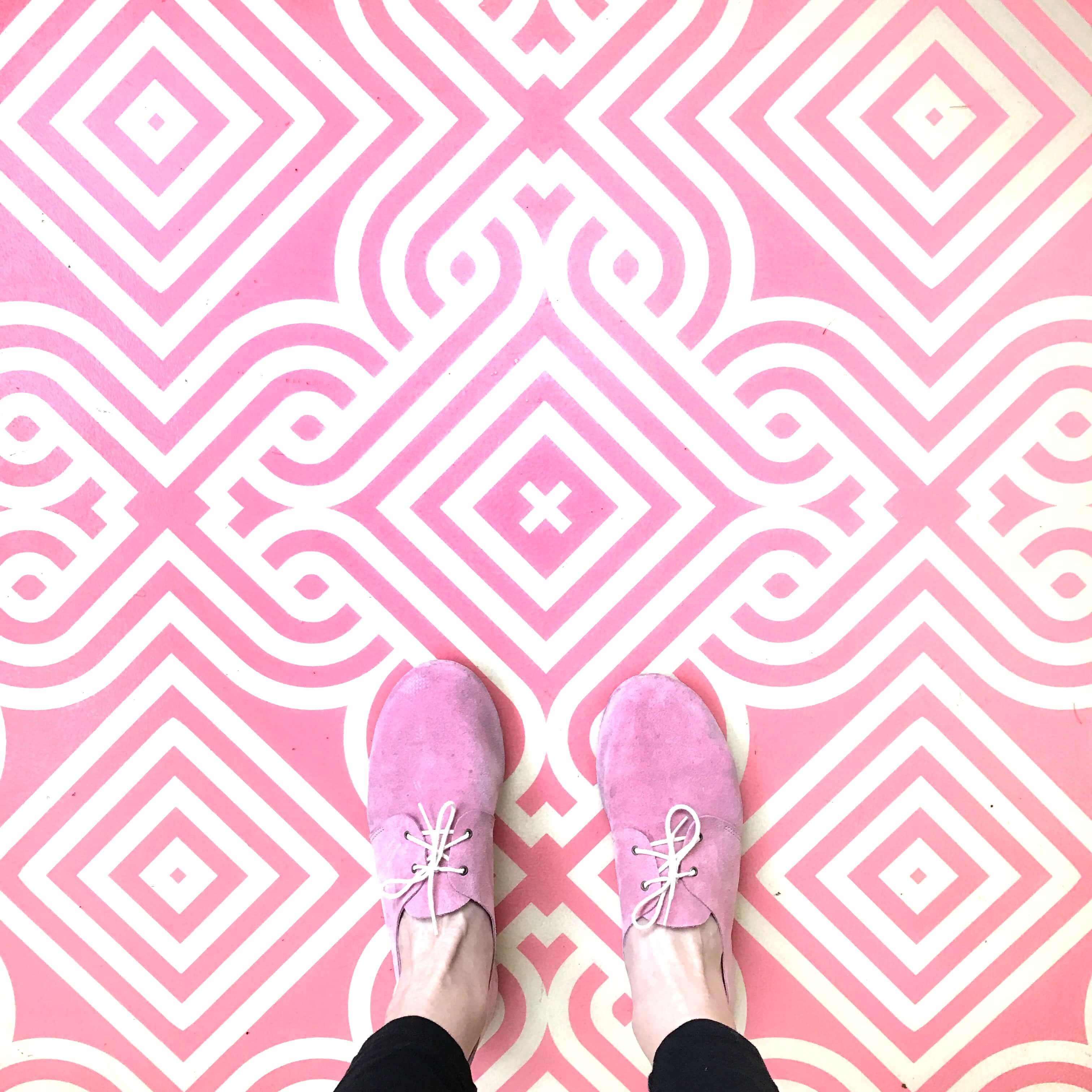 A top-down shot of pink suede lace-up shoes standing on a bold pink and white patterned tiled floor — used to illustrate a Sheldon Social article on brand voice and founder-led content.
