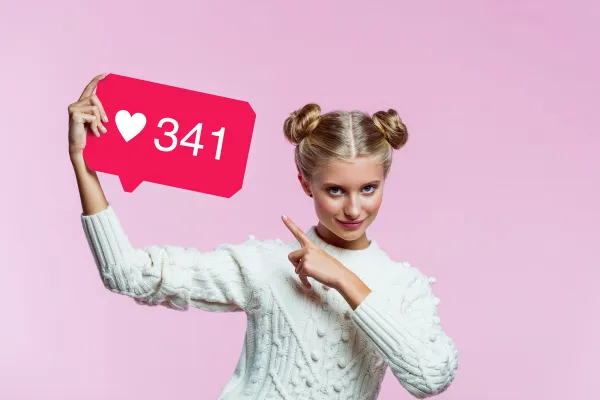 A young woman in a white jumper pointing to a social media likes notification graphic against a pink background — a decorative image used to illustrate a Sheld…A young woman in a white jumper pointing to a social media likes notification graphic against a pink background — a decorative image used to illustrate a Sheldon Social blog post on social media engagement, online visibility, and content strategy.