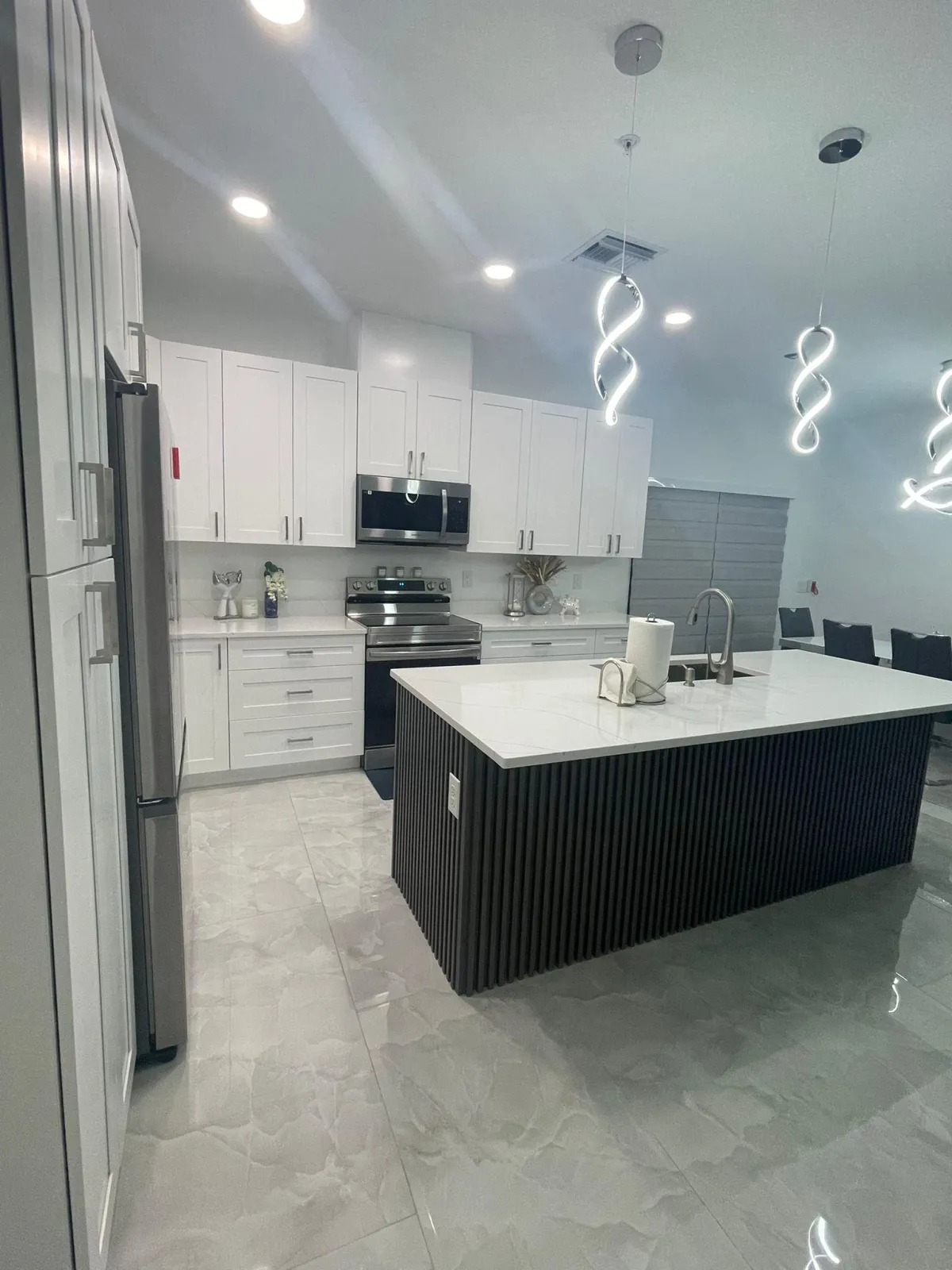 Modern white kitchen with quartz countertops, stainless steel appliances, LED pendant lights, and glossy marble-look tile flooring.