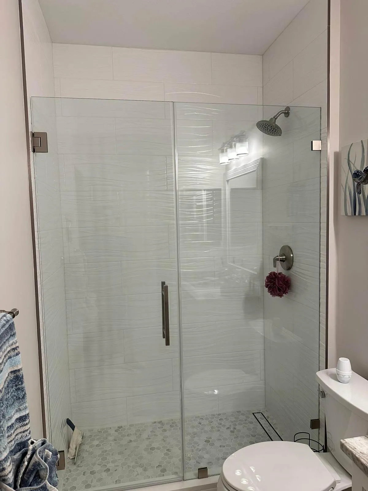 Modern bathroom shower remodel with frameless glass doors, wave-textured white wall tiles, and a hexagon tile floor with linear drain.