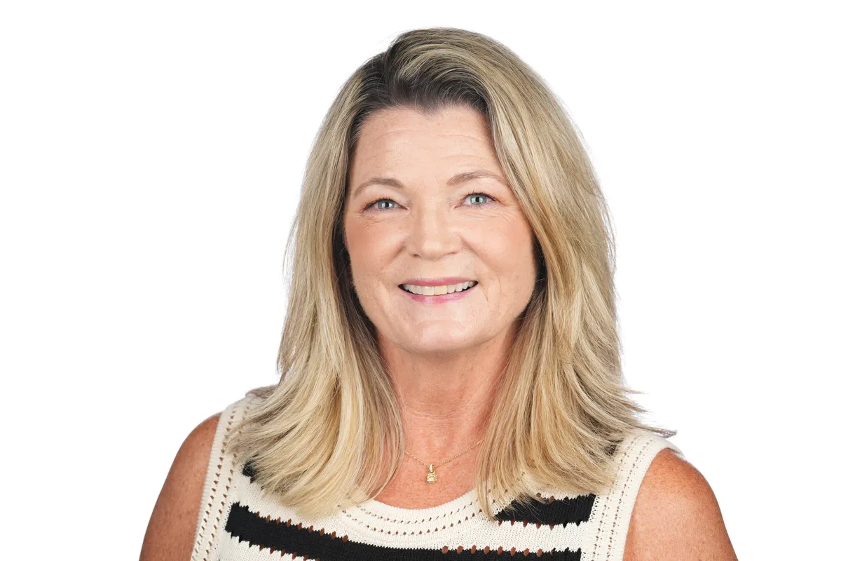Coach Kelly, a woman in her early 40s with curly hair, smiling warmly, seated at a sunlit desk with a notebook and mug, exuding approachability and encouragement in a bright, inviting workspace.