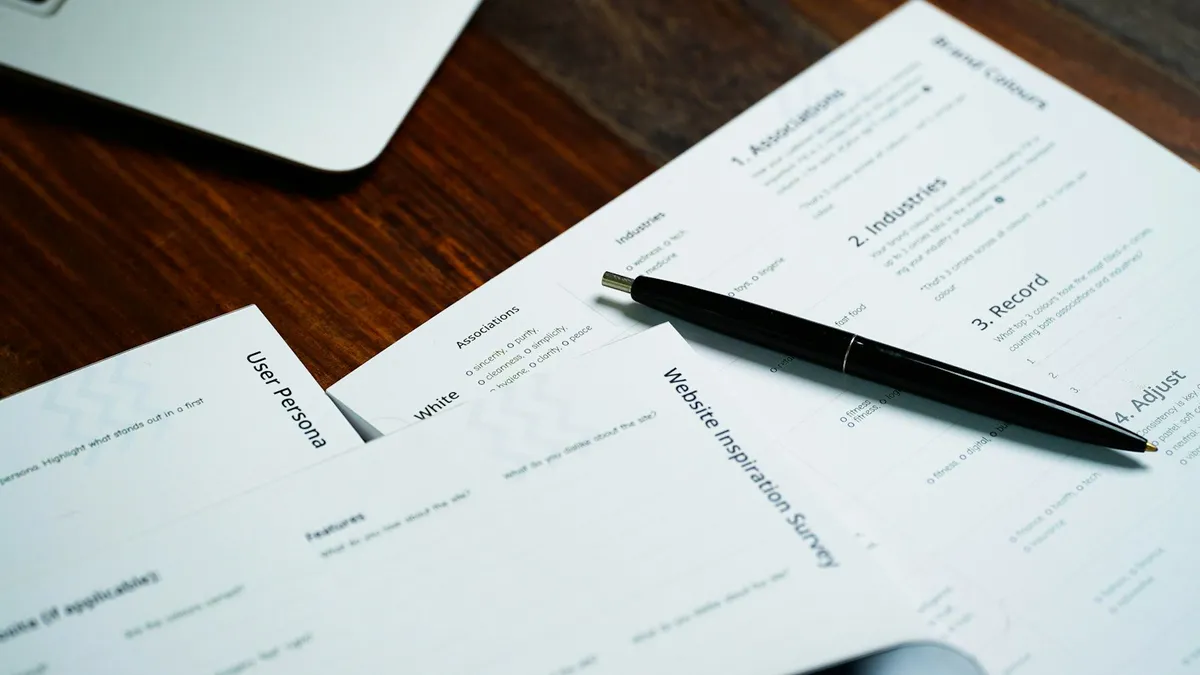 Printed paper forms spread across a wooden desk with a ballpoint pen resting on one of them