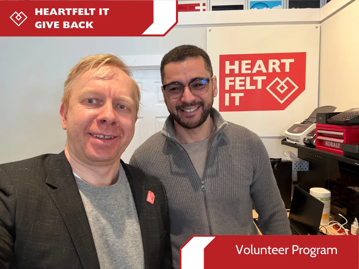 A marketing graphic for a Heartfelt IT initiative. The photo features a close-up of two smiling men in a branded workshop. One man is wearing a blazer with a small red Heartfelt IT heart pin. The background includes a large logo with the text "HEART FELT IT" and the white heart-diamond symbol, along with a stack of technical equipment and a Kobalt-branded toolbox. Red and white banners at the top-left and bottom-right read "HEARTFELT IT GIVES BACK" and "Volunteer Program," respectively.