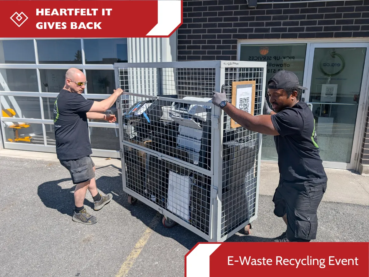 wo workers in black t-shirts moving a large metal security cage filled with donated printers and electronics outside a brick building. The image features "HEARTFELT IT GIVES BACK" branding and a red banner labeled "E-Waste Recycling Event.