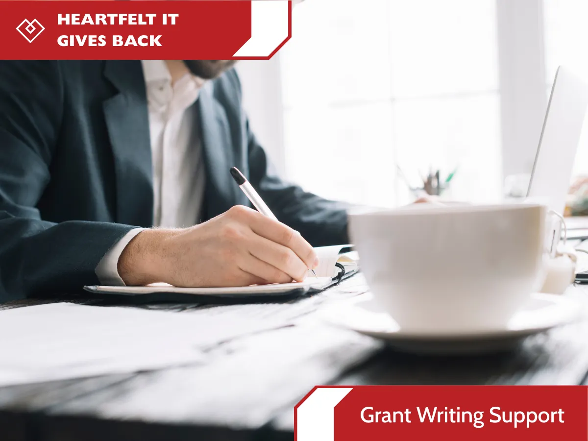 An office worker in a suit writing in a notebook on a desk next to a laptop and a coffee cup. Red banners on the top left and bottom right feature the text "HEARTFELT IT GIVES BACK" and "Grant Writing Support".