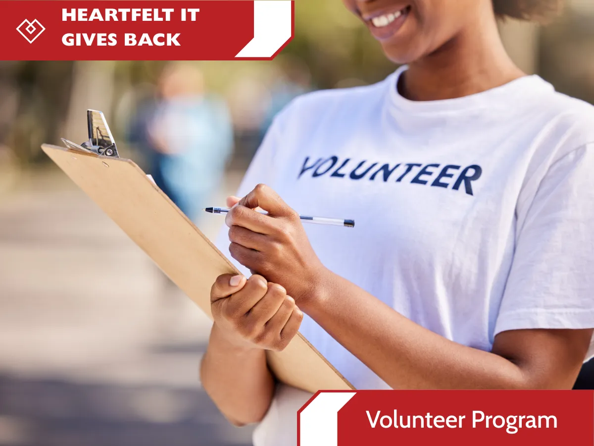 A smiling volunteer in a white t-shirt writing on a clipboard, with red Heartfelt IT Gives Back banner graphics indicating the Volunteer Program.