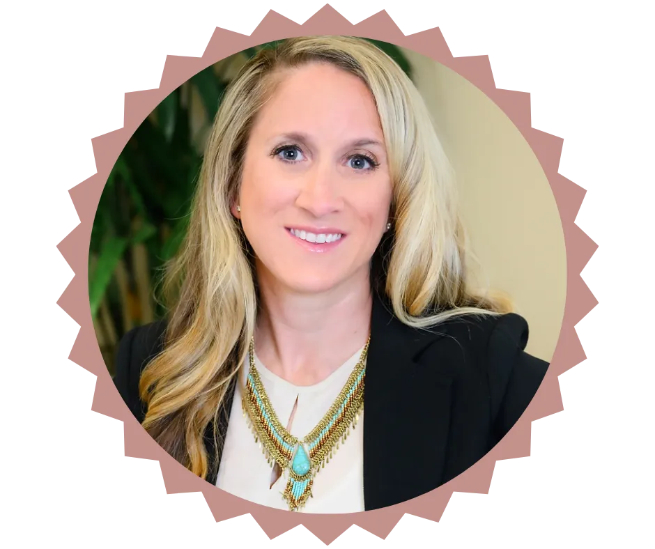  Portrait of Sarah Monceaux, founder of Unity Psychiatry, smiling warmly. She has long blonde hair, is wearing a black blazer and a layered necklace with turquoise accents, and is positioned against a soft background with greenery.