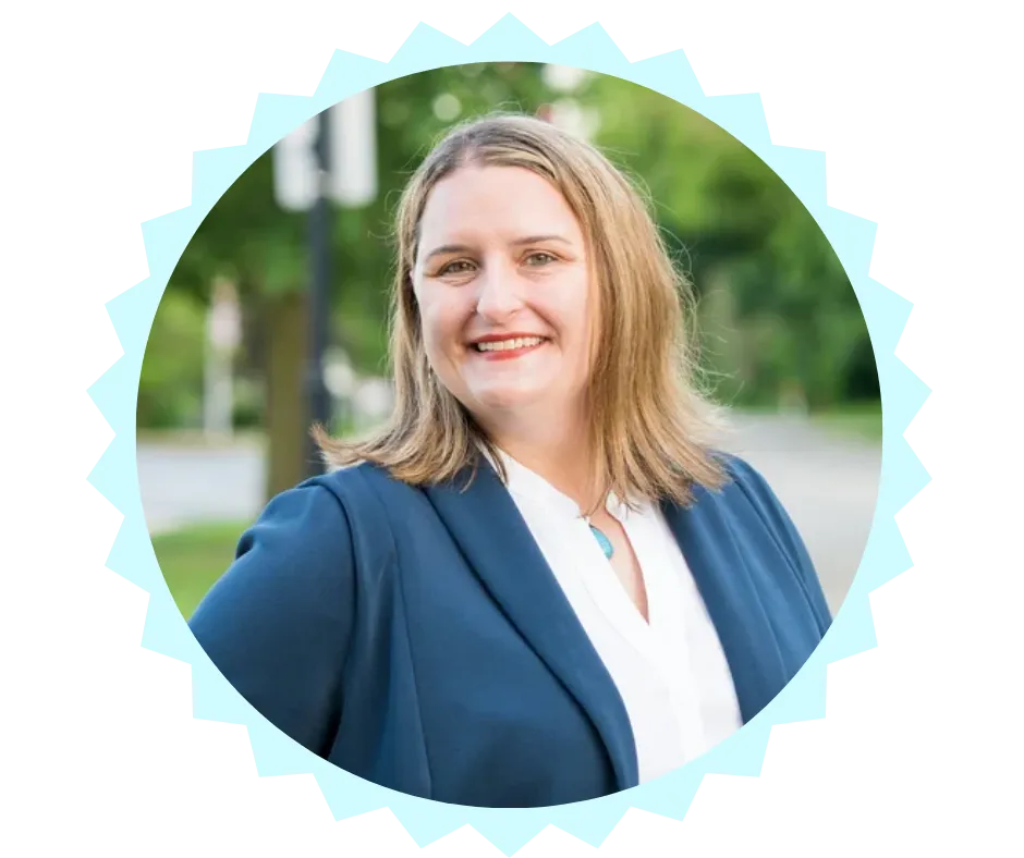 Portrait of Sarah Harrison, candidate for Wisconsin State Assembly, smiling confidently outdoors. She is wearing a navy blazer over a white blouse with a turquoise pendant, standing against a blurred background of greenery and a street.