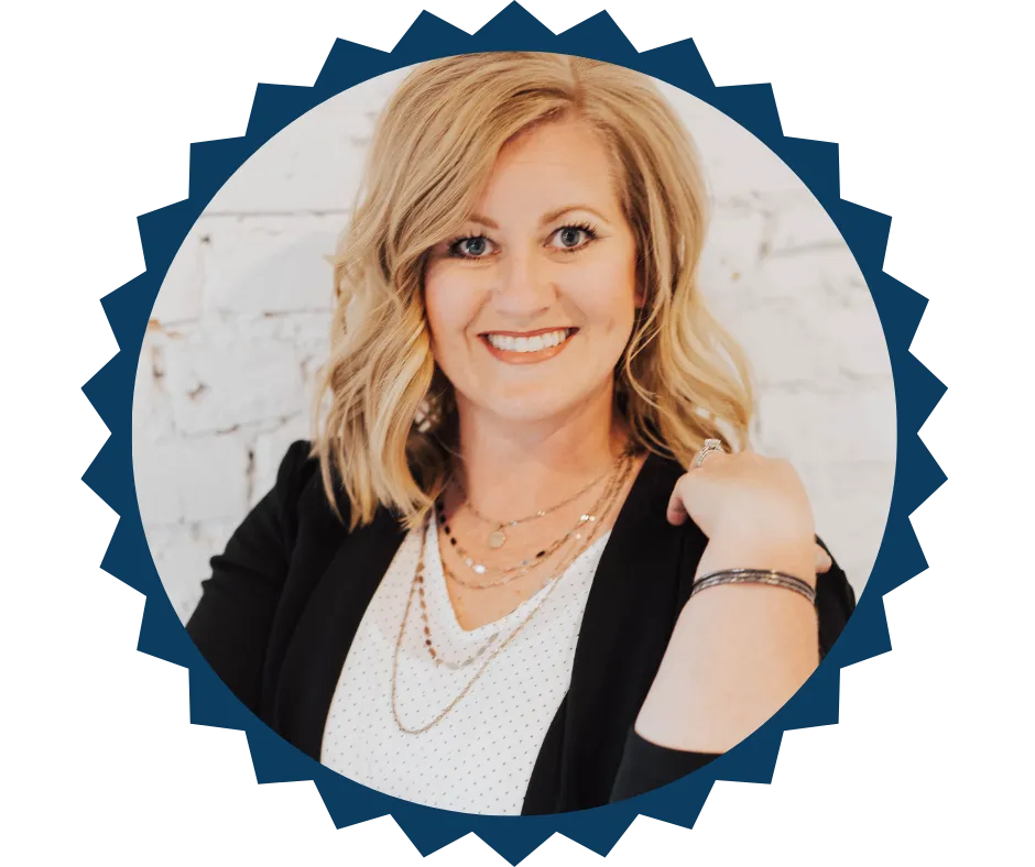 Portrait of Kristen Kippen, owner of Strategic Tax Planning, smiling confidently. She has shoulder-length blonde hair and is wearing a black blazer over a white blouse, accessorized with layered necklaces and bracelets, set against a white brick background.