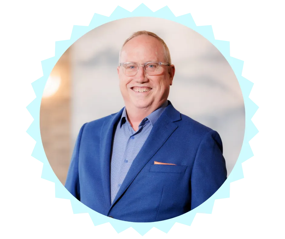 Portrait of Chris Pruitt, a financial and retirement advisor, smiling warmly. He is wearing a blue blazer with a light blue shirt and an orange pocket square, standing against a softly blurred professional background.