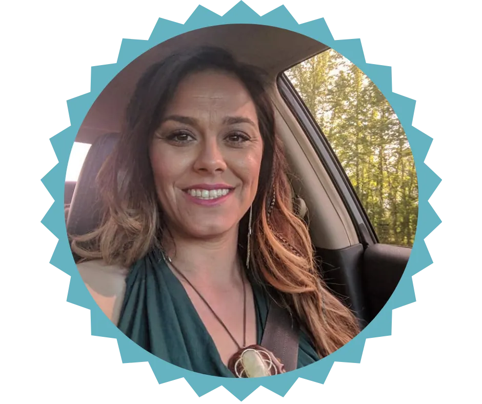 "Portrait of Kristian, owner of Euphoria Botanicals, smiling inside a car. She has long brown hair with blonde highlights, is wearing a green top, and a unique pendant necklace, with sunlight and trees visible through the window."