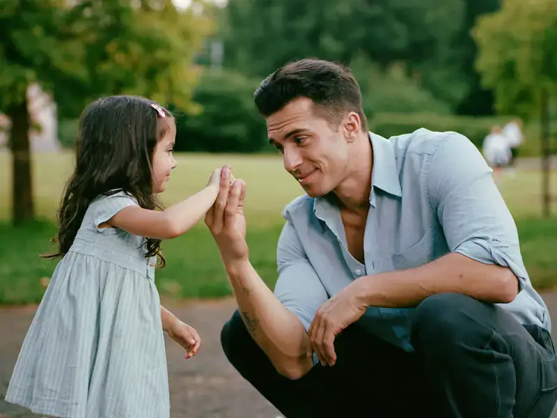 A man crouching down smiling at a young girl holding up her hands in a playful manner, outdoors with trees in the background.