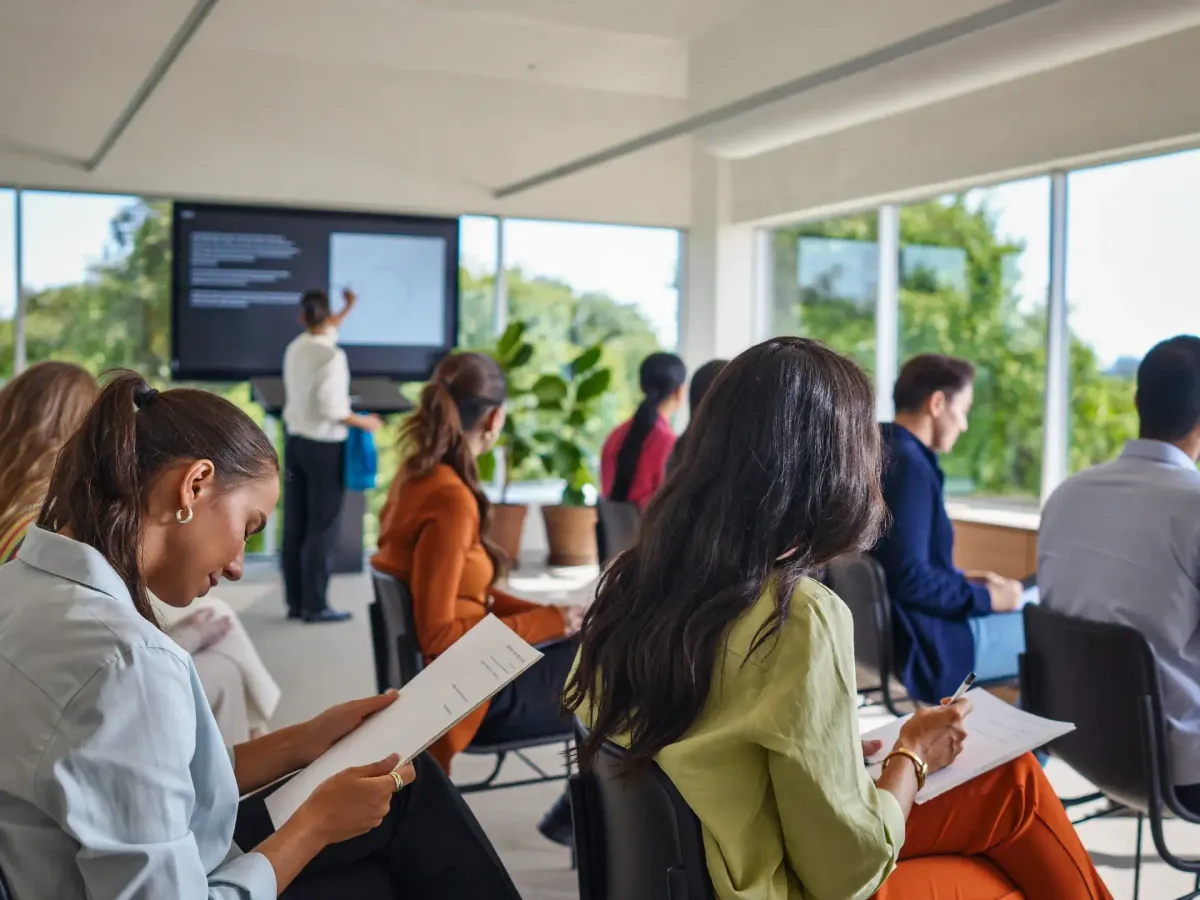 A group of professionals sitting around tables in a modern training room, listening to a presentation with a screen in the background.
