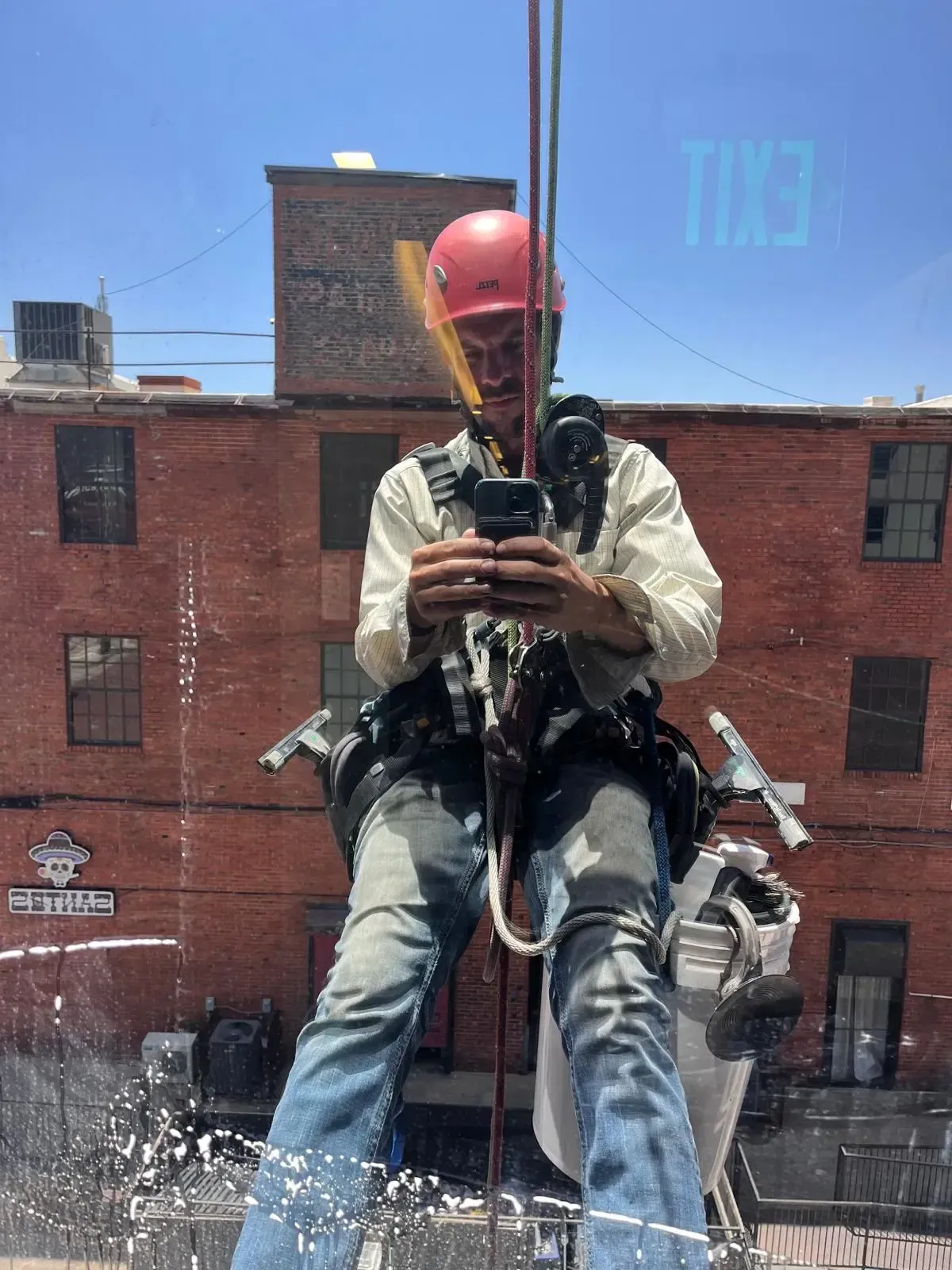 Professional window cleaner in safety harness and hard hat using pressure washing equipment on high-rise building - SudsMonkey Window Cleaning Oklahoma City services