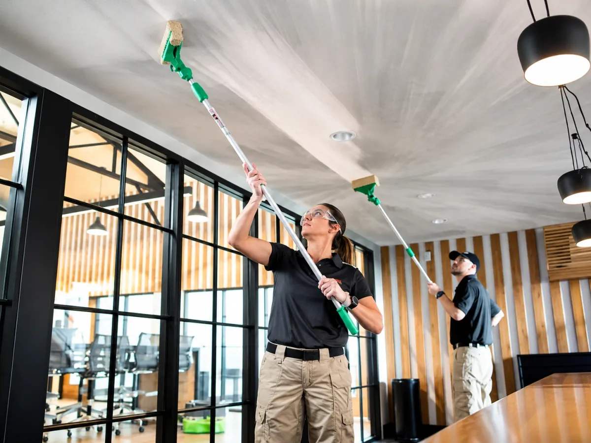 Professional cleaners removing dust from high ceilings using extension poles in a modern commercial office.