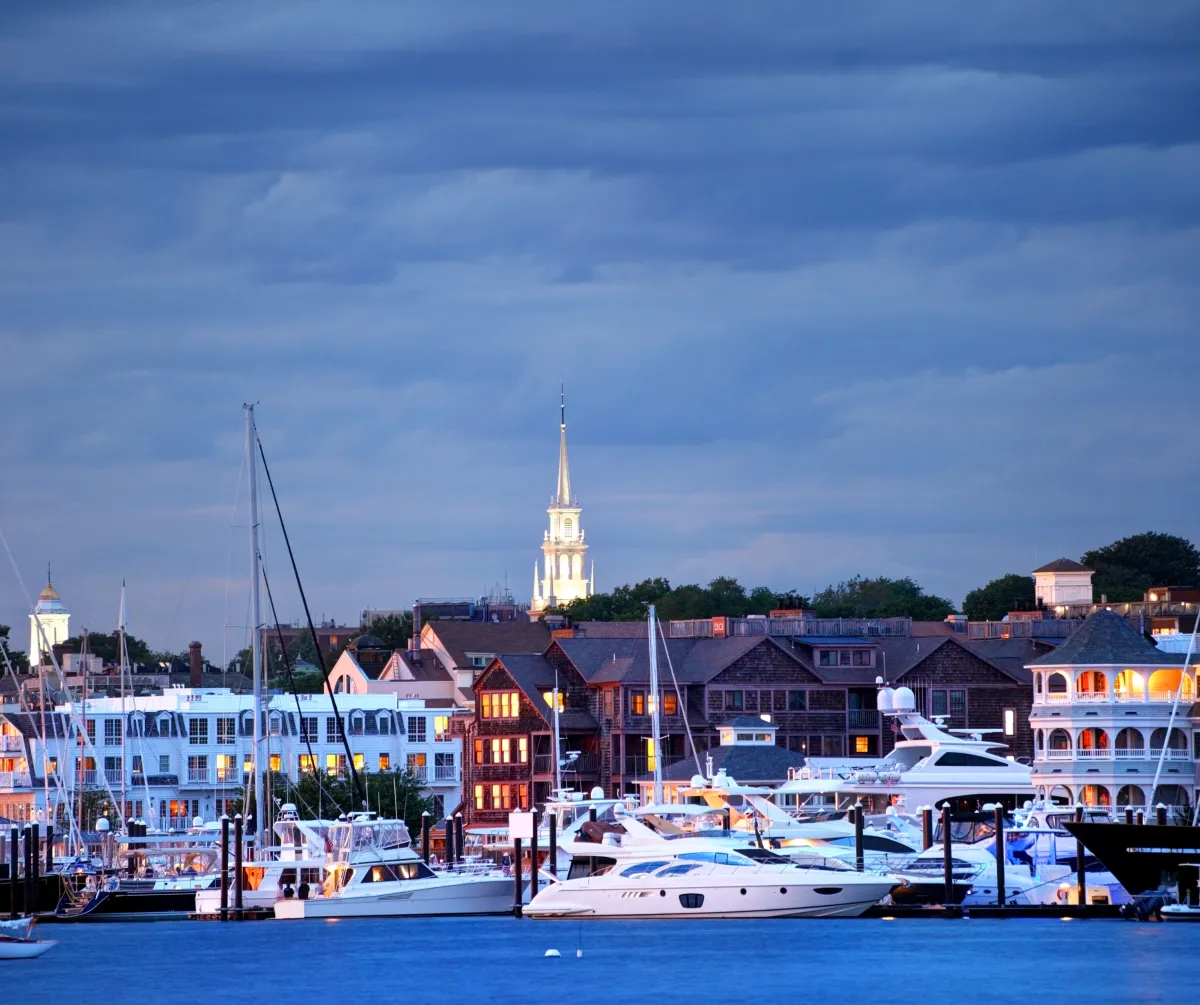 Scenic waterfront view of Newport, Rhode Island at dusk, with yachts docked in the marina and historic church steeple in the background.