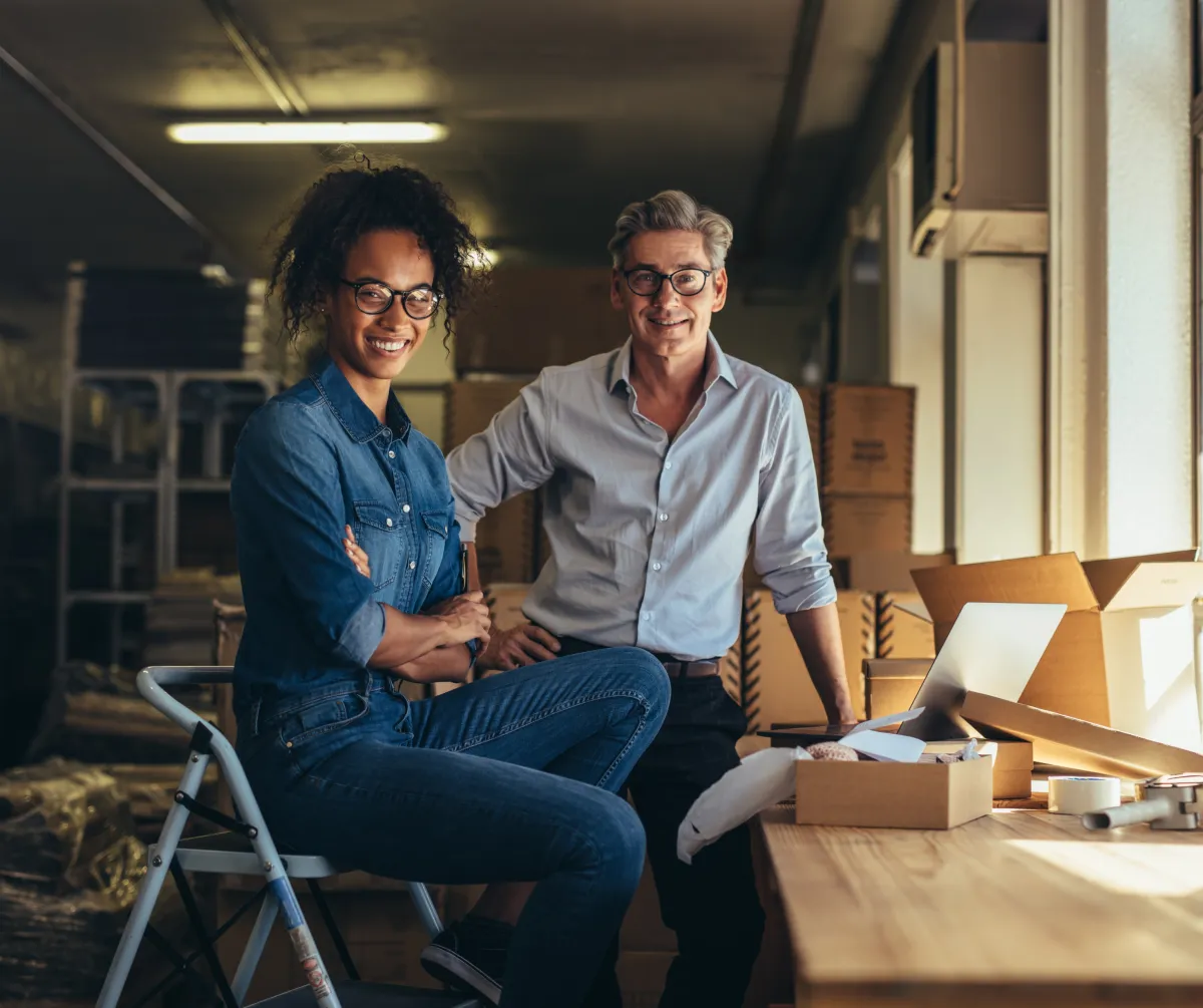 Two Newport small business owners smiling in a workshop filled with packaging supplies, symbolizing the hardworking local entrepreneurs supported by Rhode Island AI.