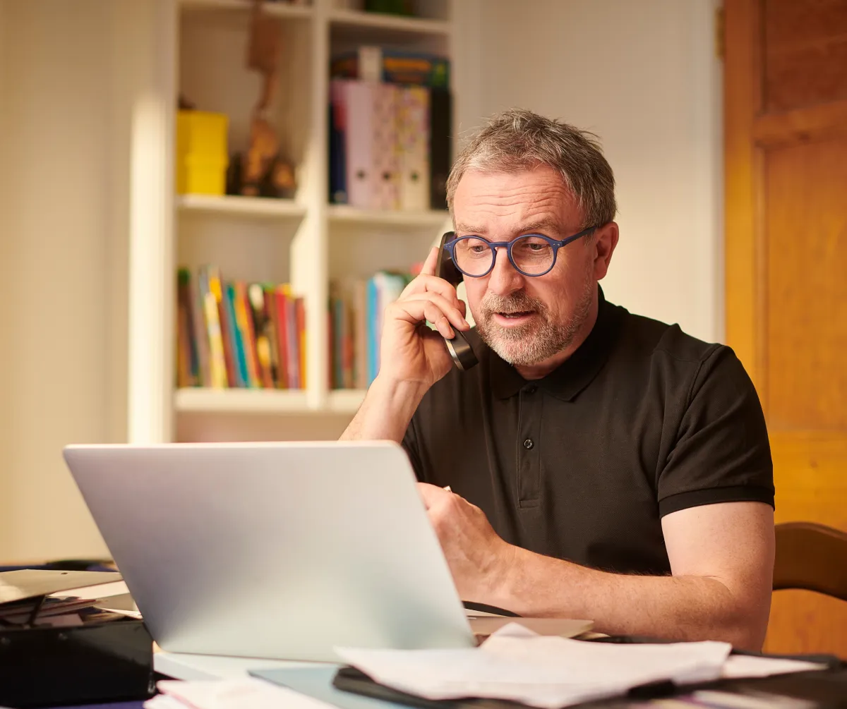 Mature man in a home office wearing glasses, speaking on the phone while using a laptop, representing a local Narragansett business owner managing client communication remotely.