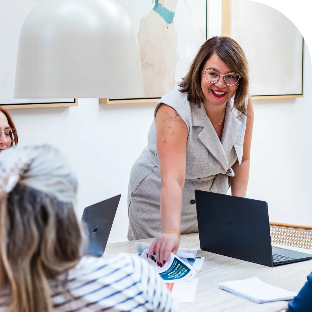 Angela Hodgson standing at a table in a modern meeting room, smiling as she hands printed materials to clients seated with laptops during a collaborative work session.