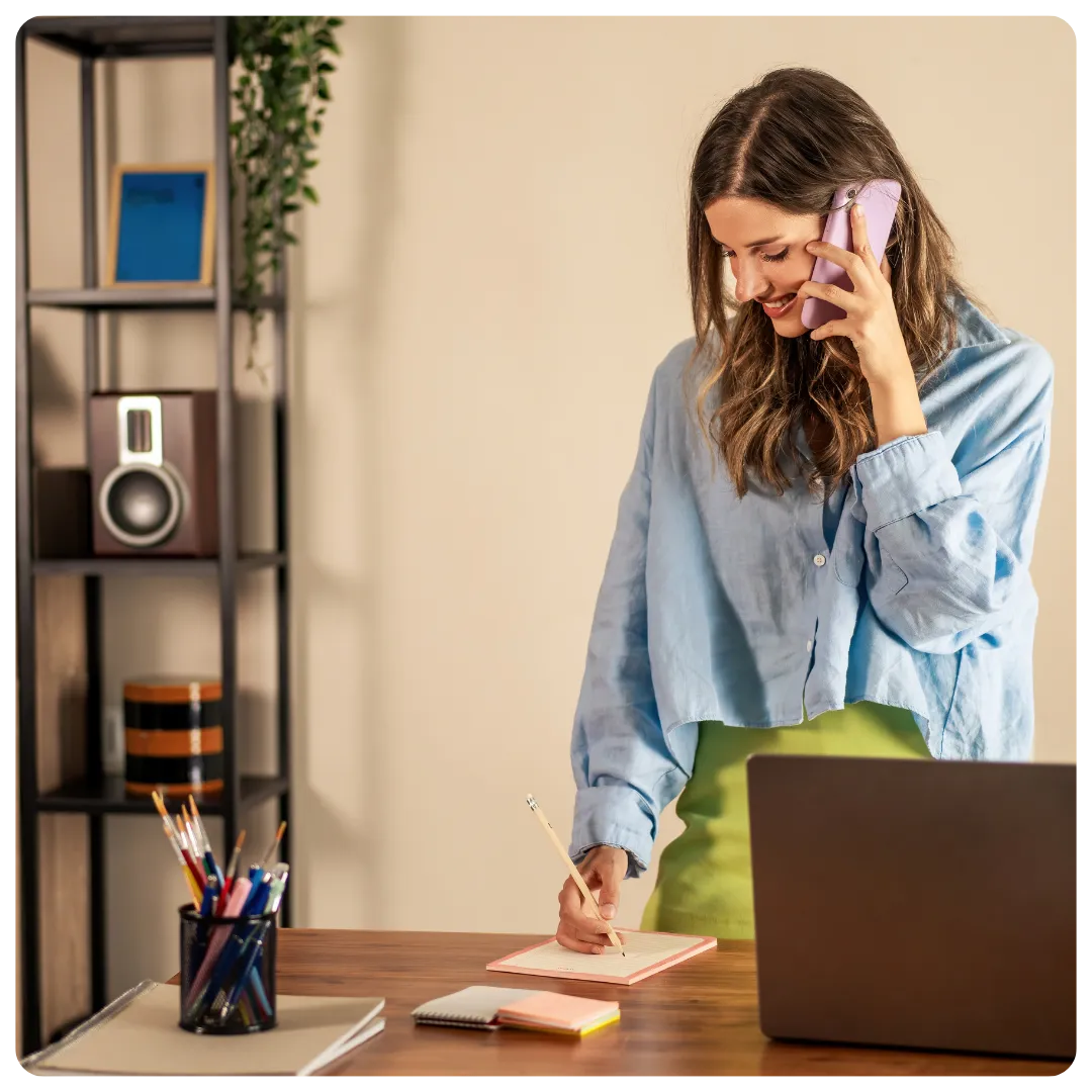 A woman standing at a desk, smiling while talking on a mobile phone and writing notes in a notebook beside an open laptop in a home office.