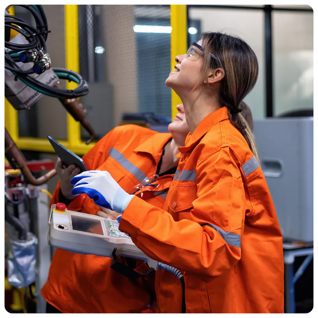 Two factory workers in orange high-visibility coveralls and safety glasses operate industrial robotic equipment. One worker holds a control panel while looking up at the machinery, and the other stands behind holding a tablet in a modern manufacturing environment.