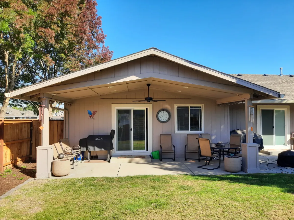 Patio area of a modern home featuring a covered porch, outdoor seating, and a grill, surrounded by a well-maintained lawn and trees, illustrating outdoor living space potential for residential construction projects.
