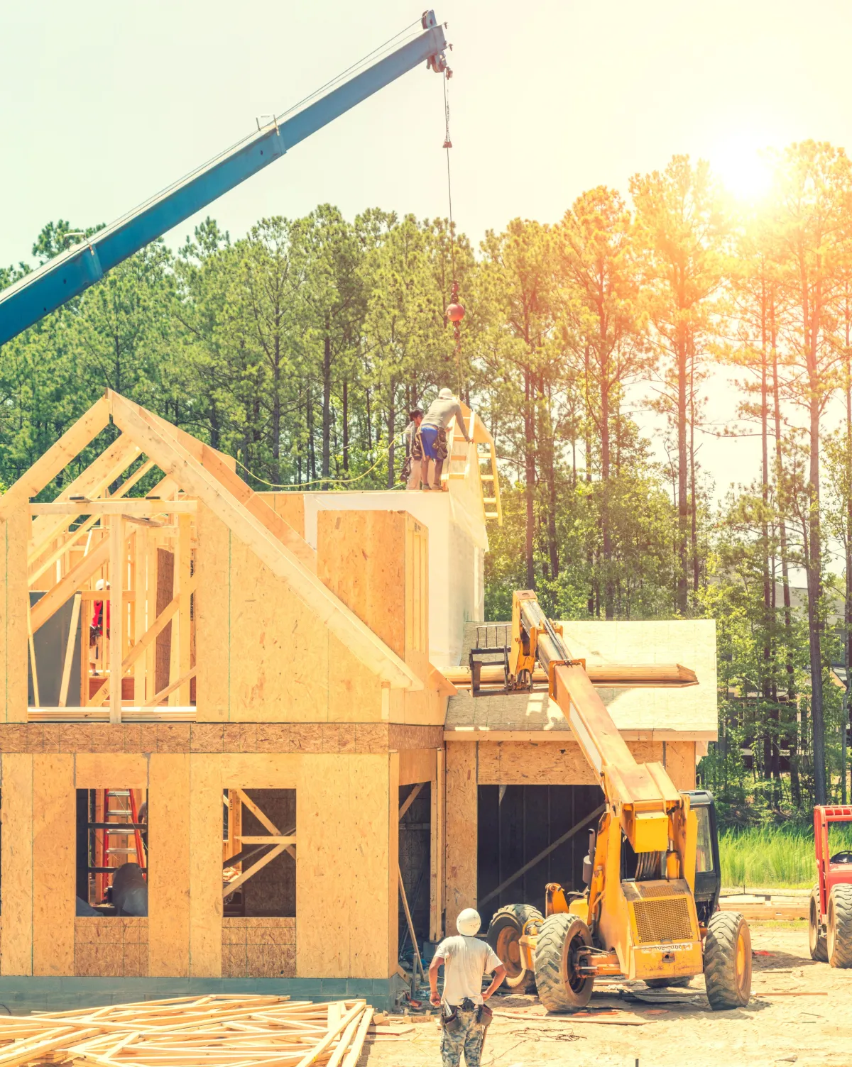 Construction crew framing a new residential home with crane and heavy equipment on an active building site surrounded by trees.