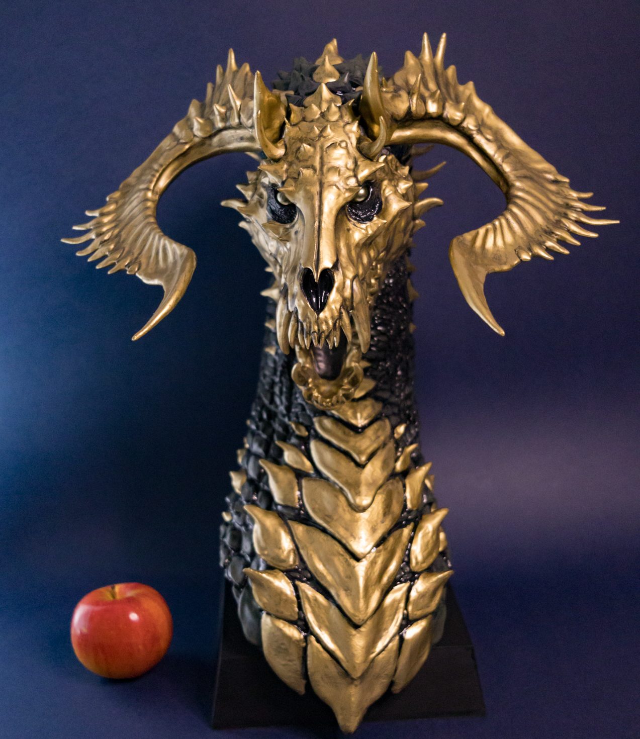 Vor’Gash Skulptur von Winton Afrić – gold-schwarze Drachenbüste mit hornartigem Schädel und strukturierter Schuppenoberfläche auf Sockel, neben rotem Apfel als Größenvergleich vor dunkelblauem Hintergrund.