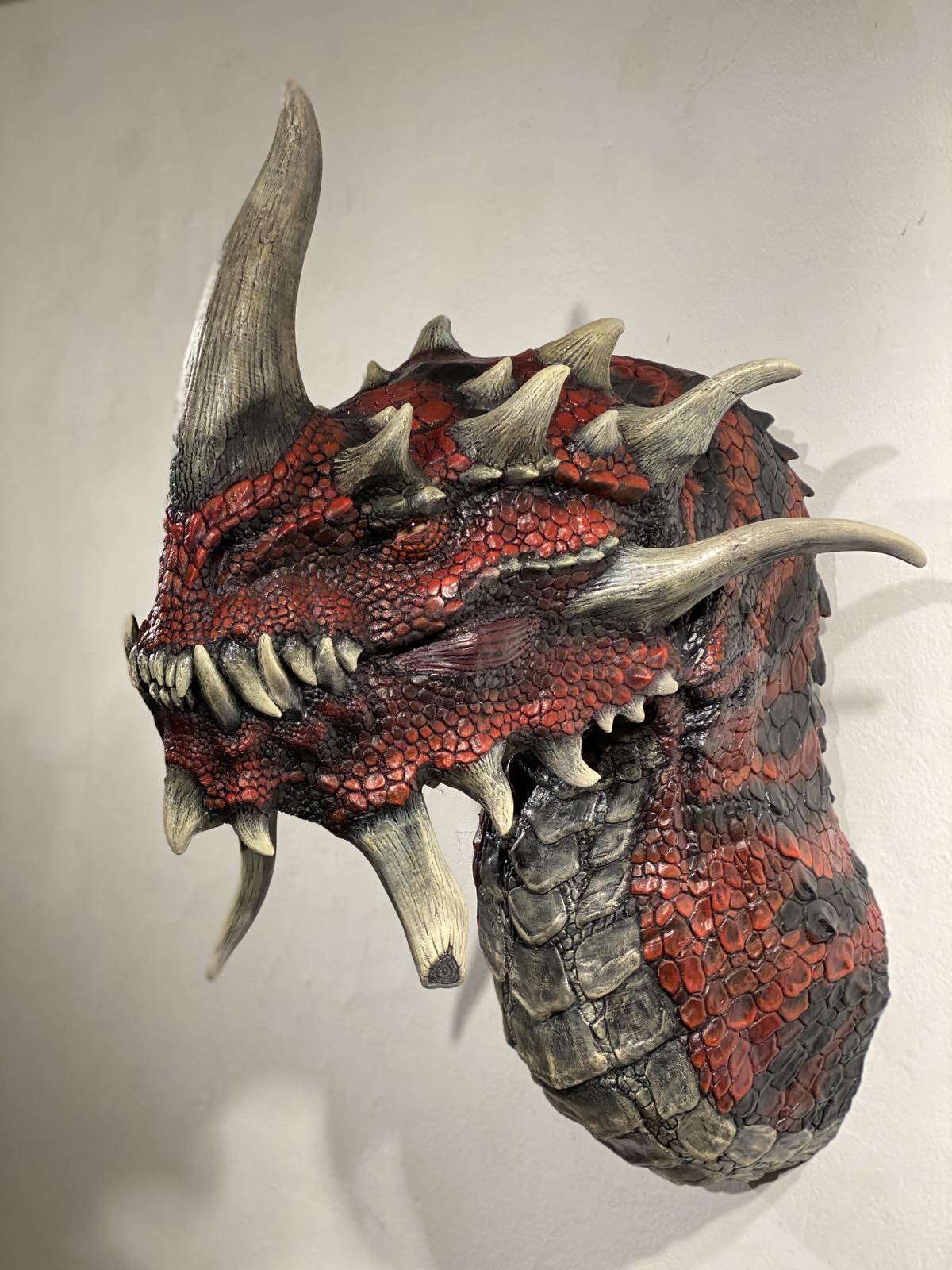 Kharzuul – Wall-Bust von Winton Afrić: rote Drachen-Wandskulptur (PLA/Kunststoff) mit massiven Hörnern, Reißzähnen und dichter Schuppenstruktur an heller Wand.