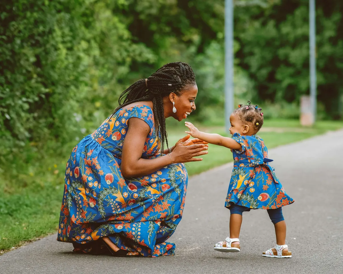 A mother kneeling on a garden path, reaching out to her toddler daughter