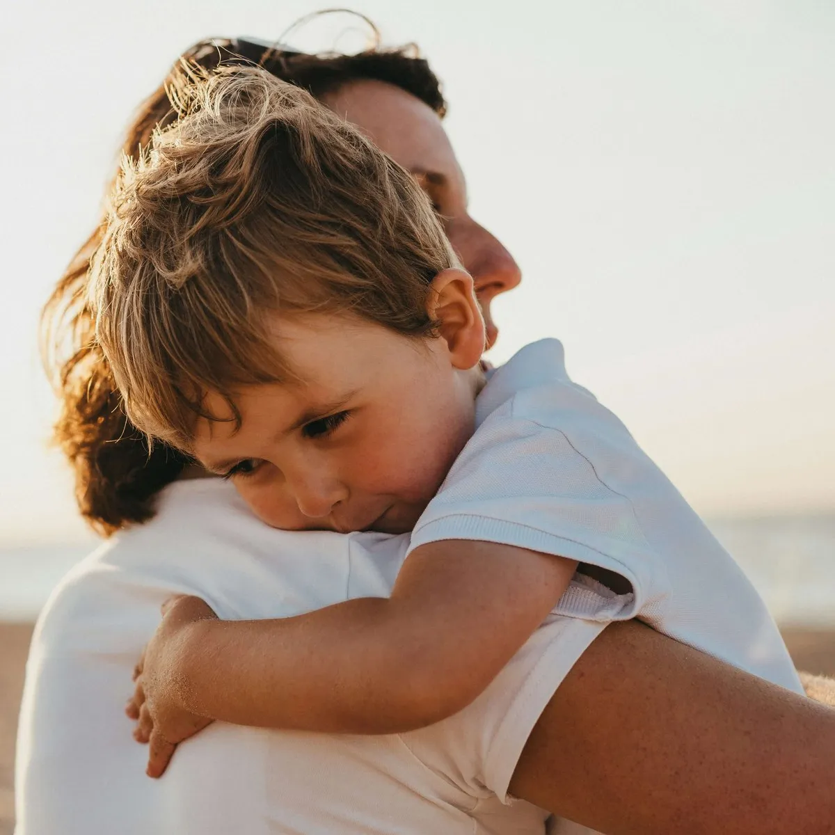 A young boy resting his head on his mother's shoulder on the beach